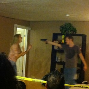 Photograph of a man pointing a gun at a frightened woman - also features three figures with their backs to the camera and are behind police tape